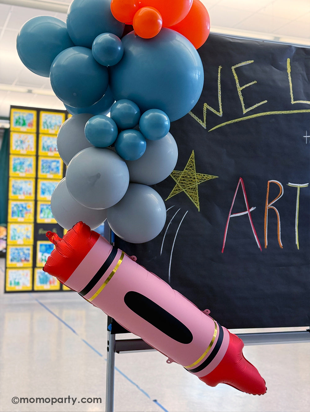 A close up shot of Momo Party's 40" x 9" red crayon shaped foil balloon along with a colorful balloon garland decoration next to a chalk board at a kid's art show in a school event. Its bright color and fun design is perfect for kid's school themed celebration, be it a back to school party, a first day of school celebration or any kids event at school.