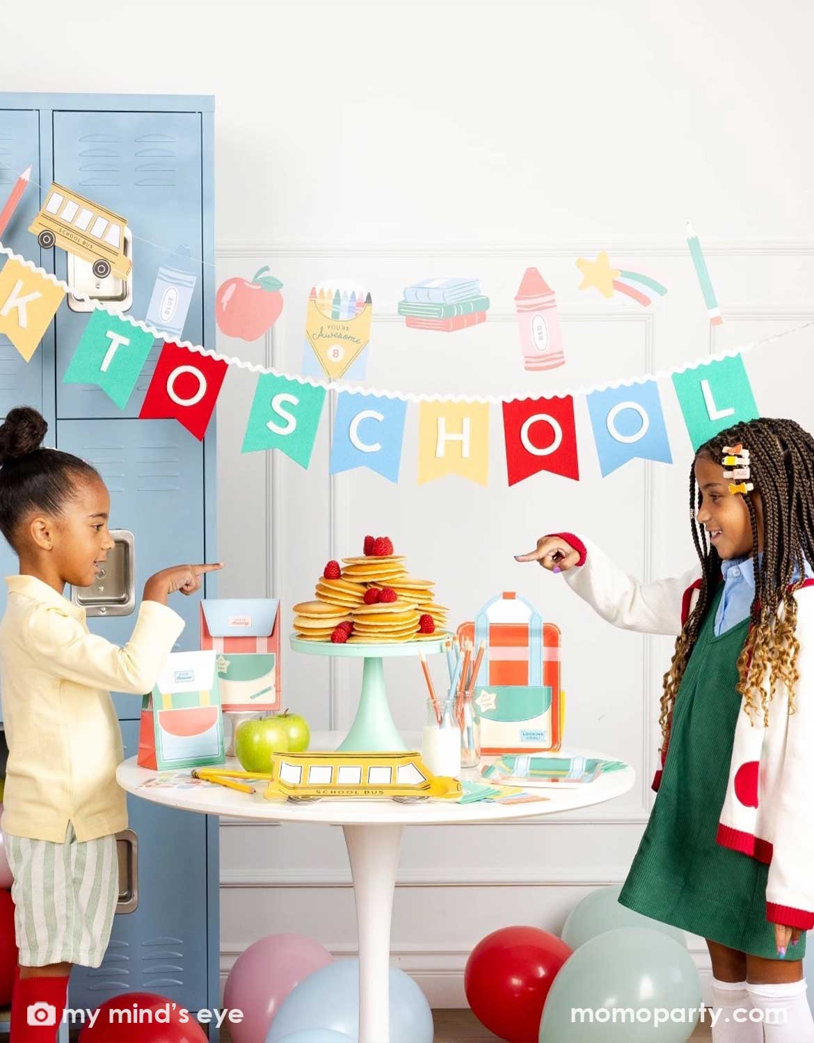 Two girls standing in a back to school party featuring various school themed party supplies and decors from Momo Party including school themed plates, treat boxes, party pennant, back to school felt garland along with school icons party banner hung above the light blue locker selves. A party table with pancakes on a cake stand next to some colorful backpack shaped treat bags. Along with colorful balloons spread across the room, this is a perfect idea for for kids first day of school celebration.