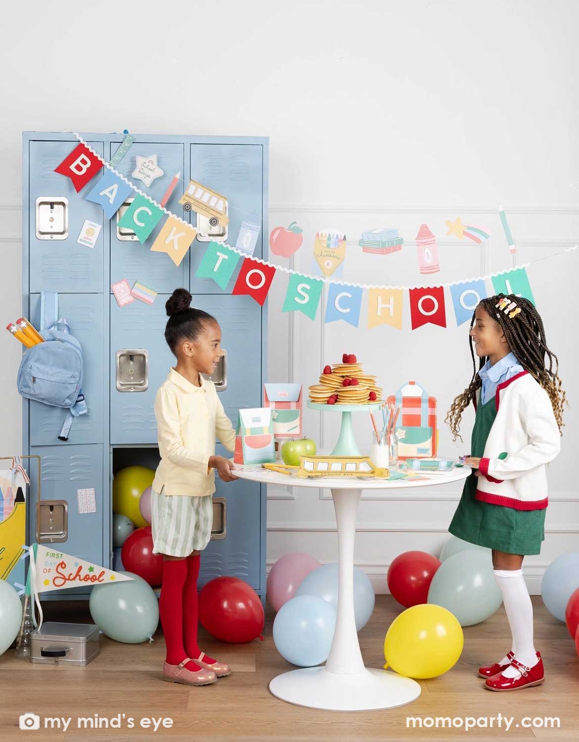 Two girls standing in a back to school party featuring various school themed party supplies and decors from Momo Party including school themed plates, treat boxes, party pennant, back to school felt garland along with school icons party banner hung above the light blue locker selves. A party table with pancakes on a cake stand next to some colorful backpack shaped treat bags. Along with colorful balloons spread across the room, this is a perfect idea for for kids first day of school celebration.