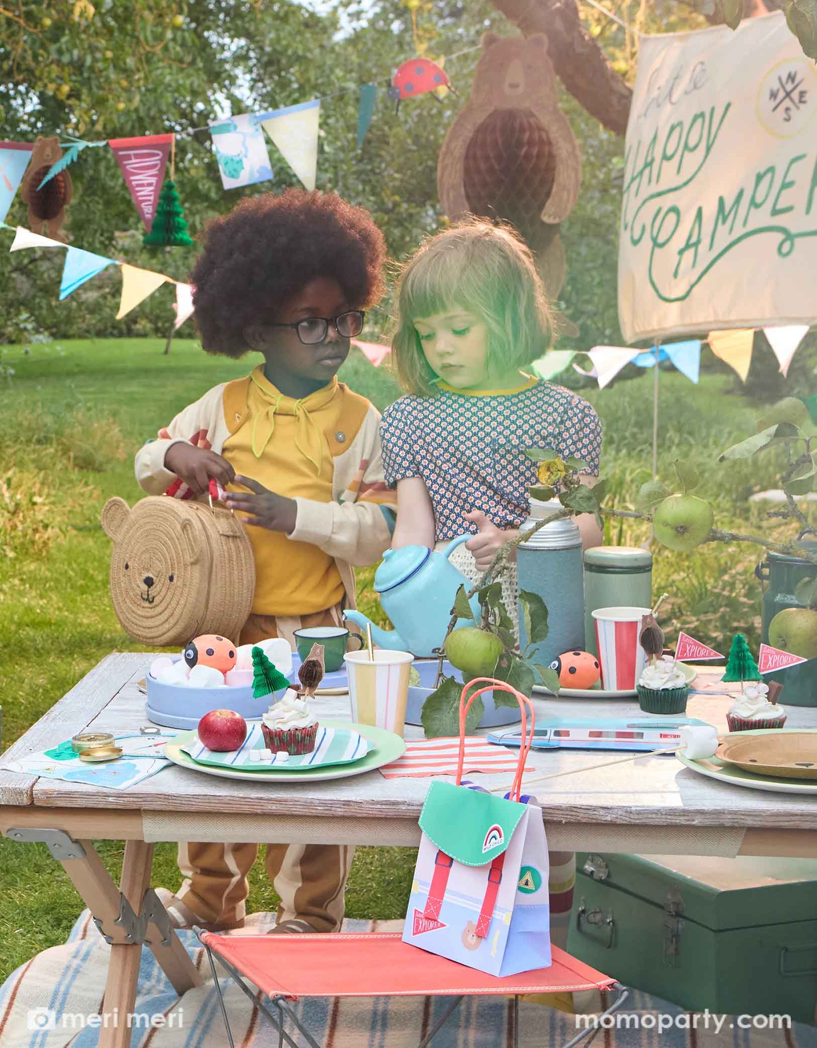 Two cute school aged girls standing next to a camping themed party table featuring multiple Meri Meri camping themed party supplies from Momo Party, including camping RV shaped plates, bear plates, compass shaped napkins, backpack shaped party bags and ladybug surprise balls. Above them hung a 10ft camping icon party garland featuring camping themed pennants and bear honeycomb. With a sign says "Happy Camper" this makes a great inspo for kid's camping themed party decorations.
