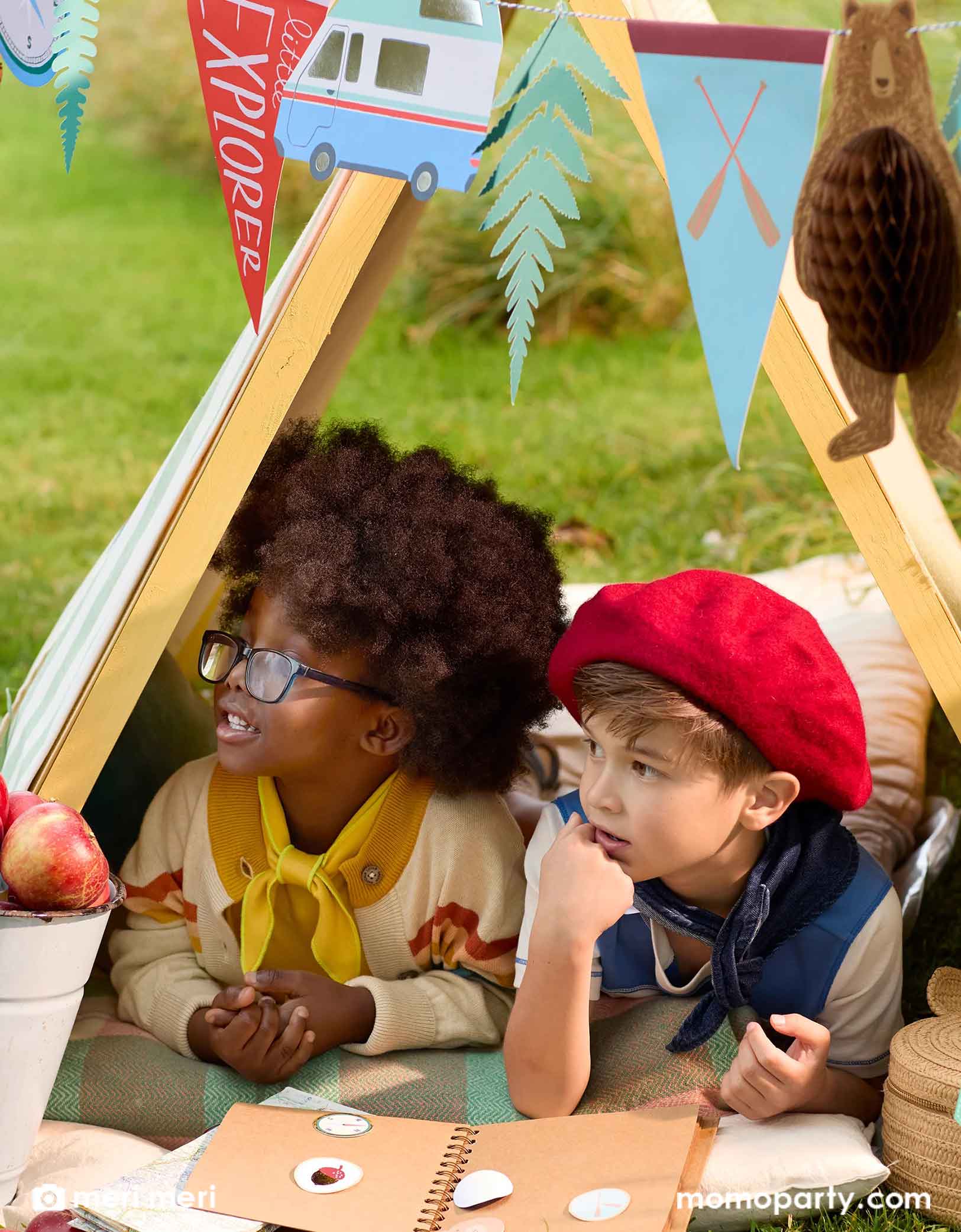 Two cute school aged kids dressed like boy scouts lying under a camping tent decorated with Momo Party's 10 ft adventure camping icon party garland featuring camping themed party pennants and bear honeycomb. Around the kids are some apples and a book of camping stickers collection.