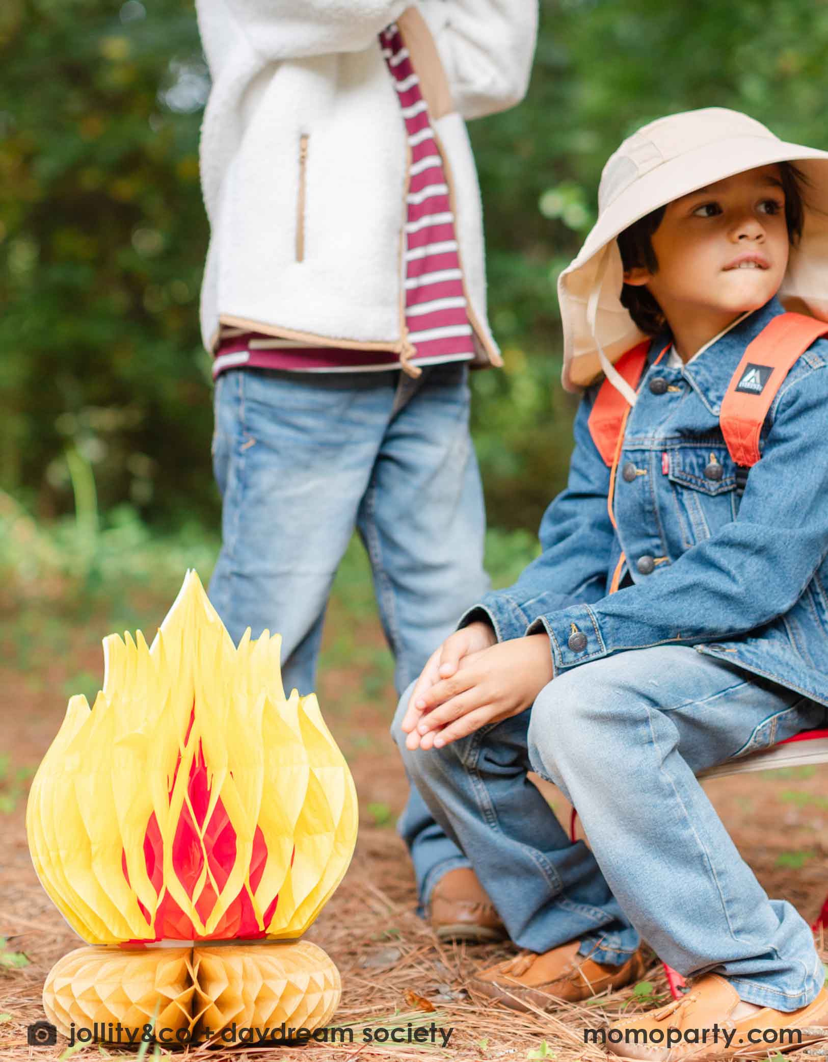 A boy sitting outdoors in his camping themed outfits, wearing a hat and backpack. with Momo Party's campfire paper honeycomb decoration on the ground.