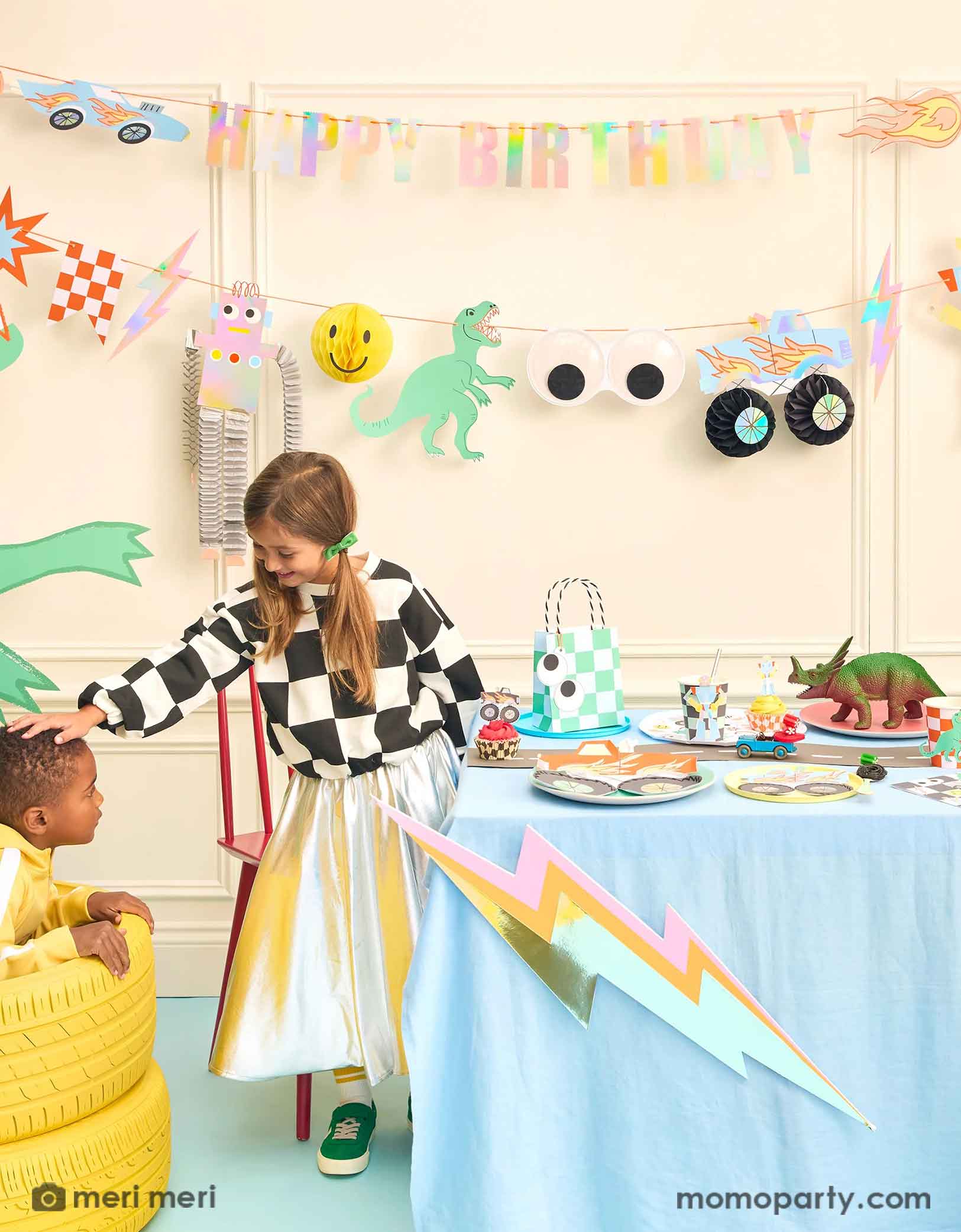 A fun themed kid's birthday party decorating set up featuring Momo Partys Momo Party's Toybox Party Collection by Meri Meri. With the retro inspired and checker board designs, featuring elements from kid's toy boxes including dinosaurs, monster trucks, race cars, googly eyes. robots and lightning bolts, these party supplies including a large party garland on the way with HAPPY BIRTHDAY message, and a table of fun and festive party tableware. Two kids playing next to the party table having a good time.