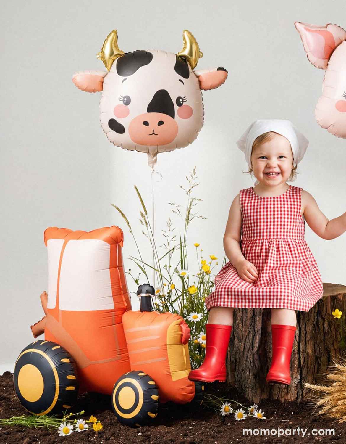 A little girl in a farm inspired red checkered dress and red boots sitting on a tree stump holding Momo Party's 23" cow-shaped foil balloon. Next to her is Momo Party's red tractor shaped foil balloon by Party Deco - a perfect inspo for kids farm themed birthdays or harvest themed celebrations!