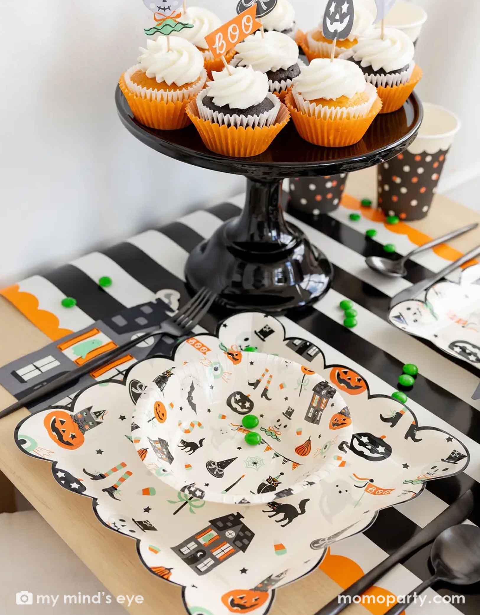 A side shot of a festive Halloween-themed table setting with Momo Party's Trick or Treat dinner plates and bowls, the haunted mansion shaped napkins and black cutlery set on a black orange and white striped table runner fro Momo Party. On the table there's a cauldron filled with green gum balls and some green candies spread across the table. A few cupcakes topped with Halloween themed toppers on a black cake stand next to it. A perfect set up for kid's friendly Halloween celebration.