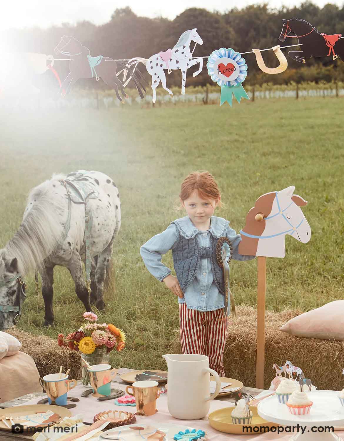 A girl in her Western themed outfits standing next to a kid's horse-themed party table setting on a lawn with horse themed party tablewares including plates, cups, cupcake kit, napkins and decorations hung above, there'a pony standing next to her in the background.