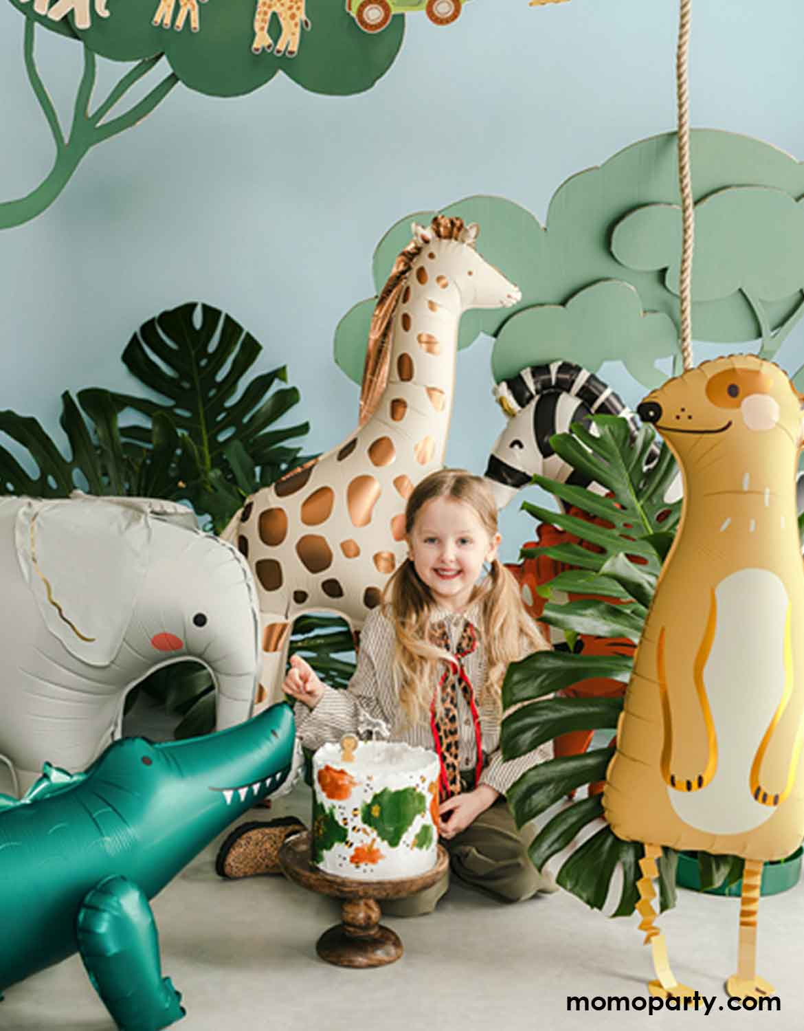 A little girl with safari adventure inspired outfits with a birthday cake surrounded by animal themed foil balloons from Momo Party including a giraffe, a zebra, an elephant, an alligator, and a meerkat in a jungle-themed setting with palm leaves around.