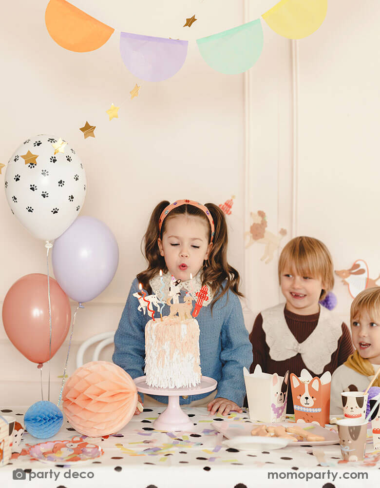 A girl blows birthday candles form a dog themed birthday cake topped with Momo Party's puppy dog cake toppers in a puppy dog themed birthday party. This party features various dog themed party supplies from Momo Party including party banners, paw balloons, corgi standing foil balloon, Dalmatian print dots tablecloth, dog shaped snack boxes, party cups and honeycomb decorations in soft pastel colors. These cute dog themed party goods make a festive touch to kid's birthday bash!