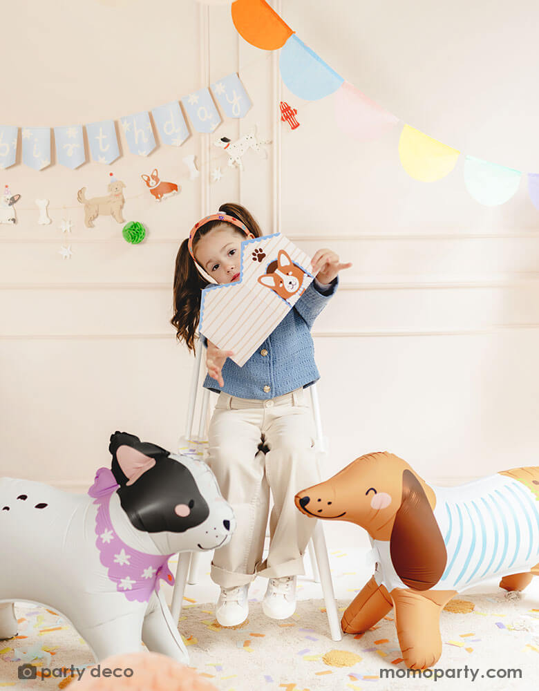 A school aged girl holding Momo Party's dog house shaped party box standing in between two dog shaped standing foil balloon in a puppy themed birthday party. On the wall there's a dog themed birthday party banner and a multi-colored bunting garland, making this a festive and fun scene for kid's birthday celebrations.
