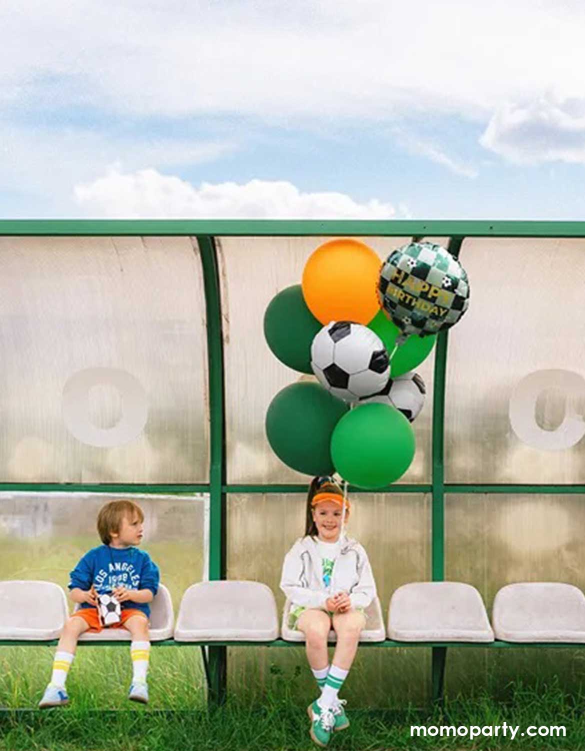 Two children sitting on a bench with soccer-themed balloon bouquet in green, orange and soccer ball shaped foil balloons from Momo Party in front of a sports facility.