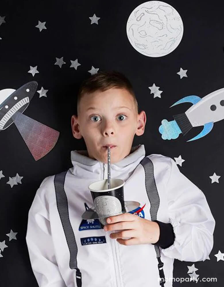 A boy in his astronaut suit holding Momo Party's 9" planet paper cups by Hooty Balloo. He's drinking from the cup with a silver foil straw, in his back the wall is decorated with space themed cutouts including rocket ships, UFOs, moon, and stars.