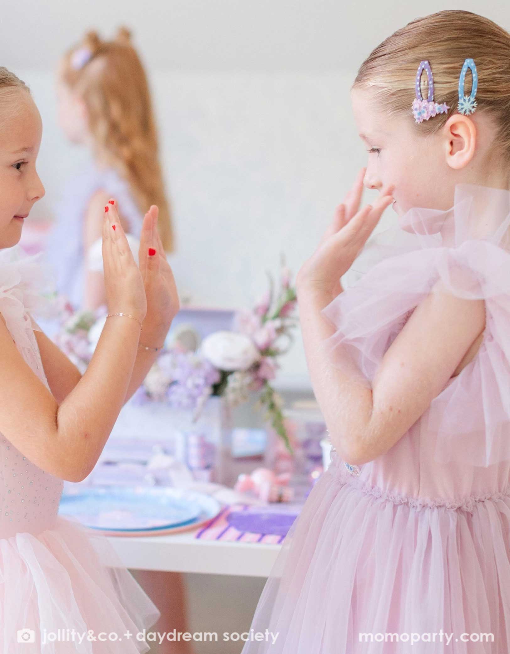 Two young girls in light pink tulle dresses with hair accessories from Momo Party featuring glittering pink and lilac Christmas tree and snowflake, clapping hands at a Christmas party with a whimsical table setting in the back.