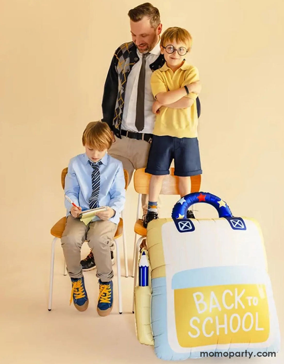 Two children in their uniform with their dad or teach standing and sitting on chairs w with Momo Party's backpack shaped foil balloon with "Back to School" text on it.