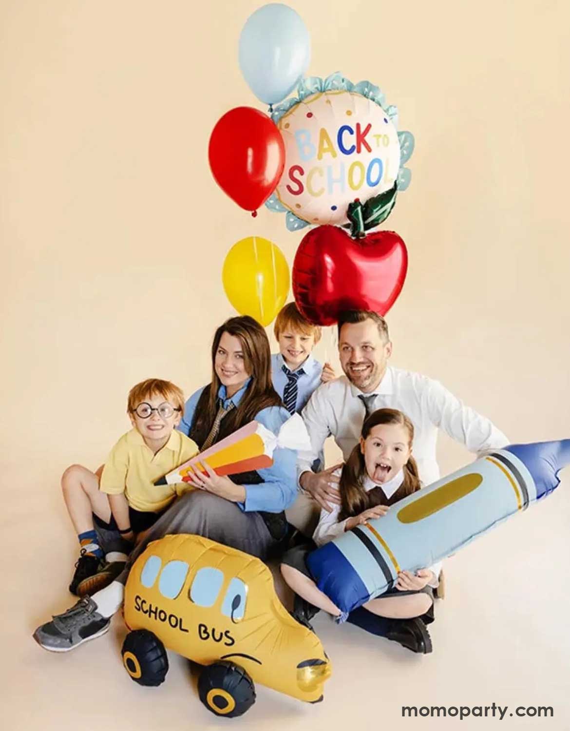 A family of five with school aged kids in their uniforms holding a balloon bouquet which features Momo Party's 20" scalloped "Back to School" foil balloon, an apple shaped foil balloon and a bunch of latex balloons in festive colors against a beige background. There are also crayon shaped foil balloon and school bus foil balloon on the ground. These festive back to school themed balloon are perfect way to celebrate your child's first day of school.