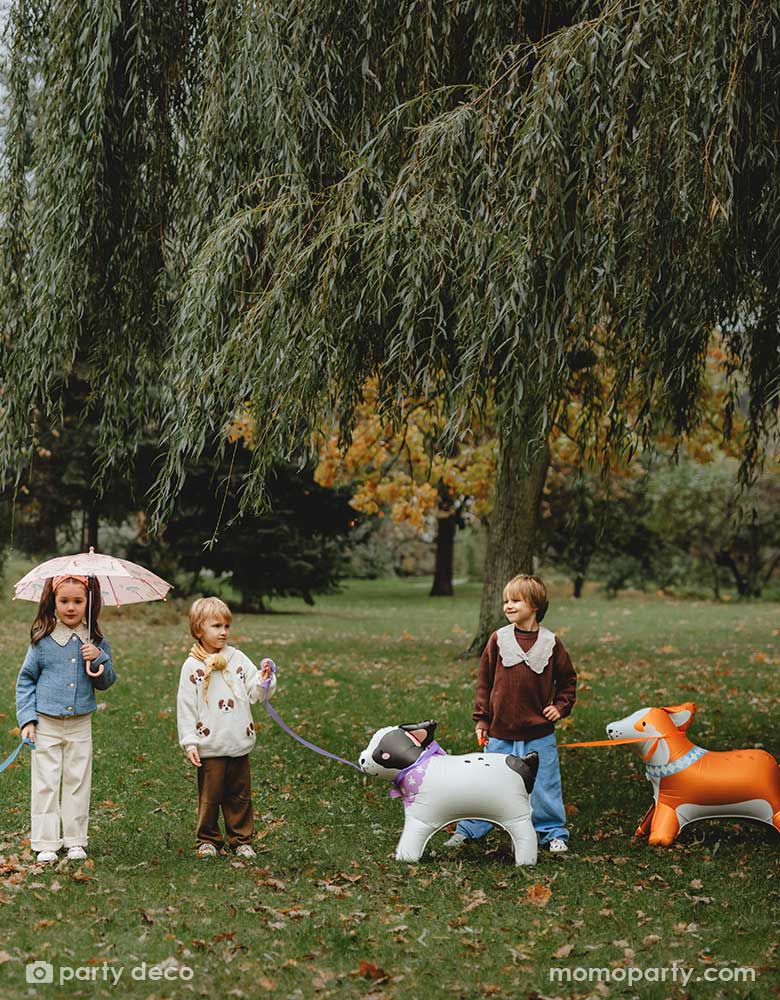 Three kids playing pretend play walking Momo Party's bull dog and Corgi standing foil balloons in a park. The girl holds an umbrela and the boys are talking.