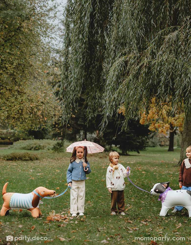 Three kids playing pretend play walking Momo Party's French bull dog and Dachshund standing foil balloons in a park. The girl holds an umbrella and the boys are talking to each other having fun in the park.