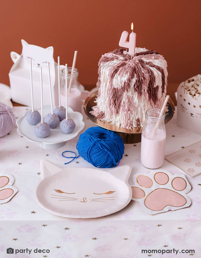 A cat themed party table featuring various cat themed party supplies from Momo Party including party plates, pink paw shaped napkins, cat shaped treat boxes filled with popcorn, small milk bottles with pale pink drink and a table of treats, snacks and a cake topped with a pink number 4 candle in pastel pink and lilac colors, making this an adorable inspo for kid's kitty cat themed birthday celebration.