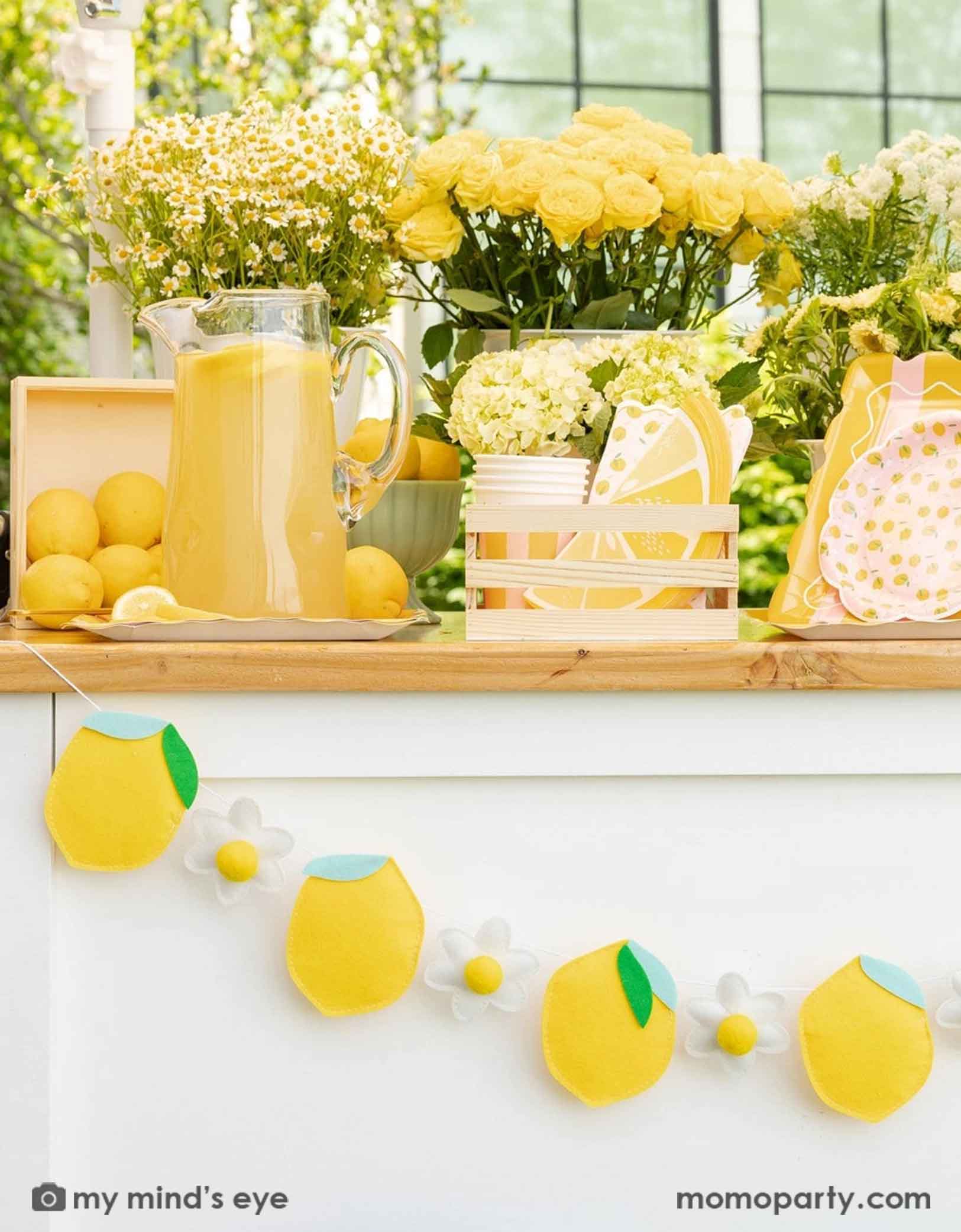 Decorative lemon garland with lemons and daisy flowers on a lemonade wooden cart with a big jar of lemonade, lemons, lemon themed party tablewares in a wooden crate and some adorable yellow and white flowers.