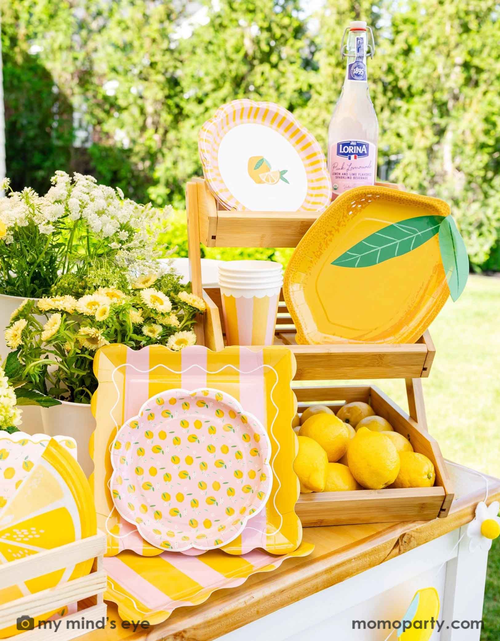 An outdoor setting with lemon themed party supplies in yellow and pink from Momo Party including lemon shaped plates, napkins and party cups on a wooden stand. With lemons and flowers around, this cute little lemon stand is perfect for a summer gathering, be it a kid's lemon themed birthday, a baby shower or a bridal shower.