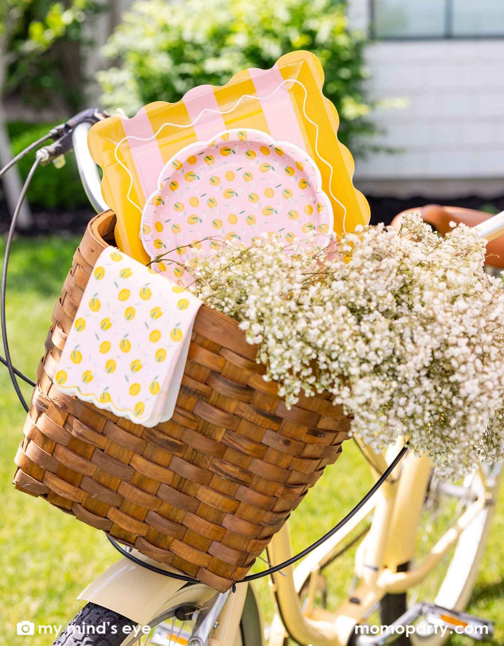 A cute bicycle basket with various of lemon themed party tablewares from Momo Party including the yellow striped square plates, lemon scattered round plates and lemon scattered dinner napkins. With flowers around, it makes an adorable scene.