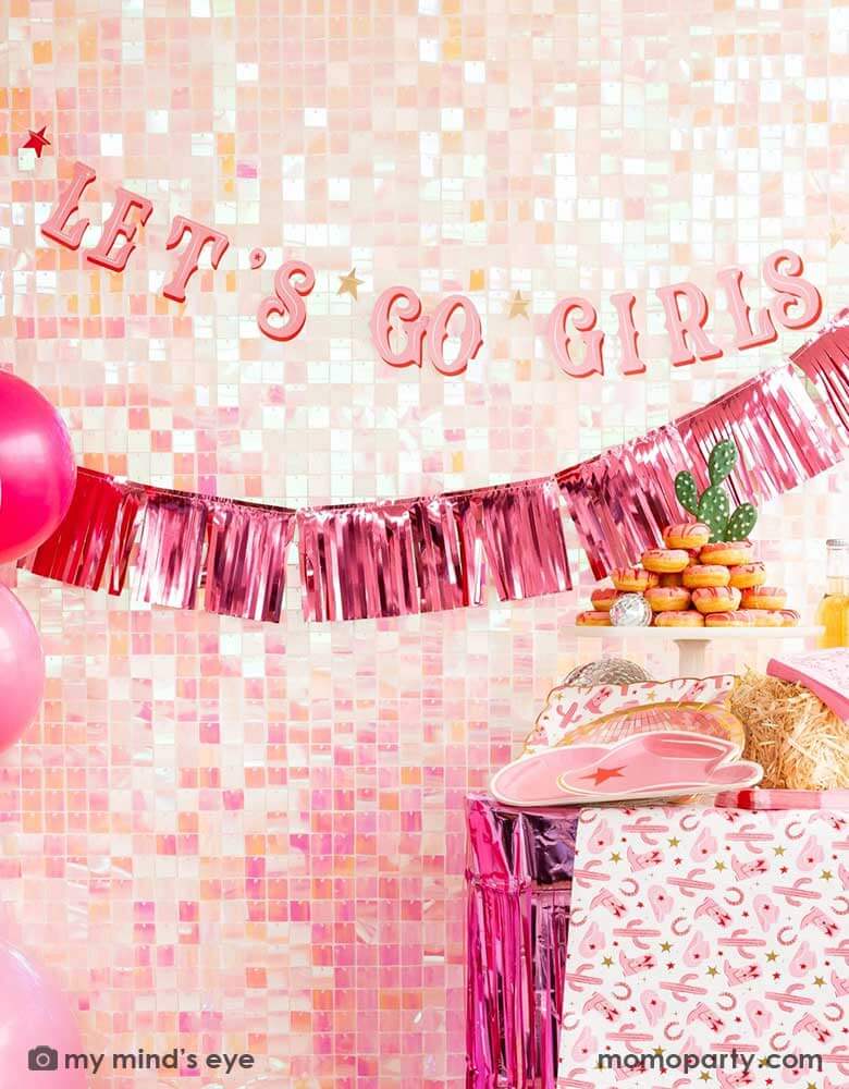A cowgirl Western themed party table features pink cowgirl themed party supplies from Momo Party including pink cowgirl boot shaped napkins, cowgirl hat shaped plates, pink disco ball shaped plates, pink bandana small napkins, yeehaw party cups and cowgirl pattern paper plates and table runner by My Mind's Eye. In the back there's an iridescent backdrop adorned with Let's Go Girls pink foil fringe banners, along with a pink donut tower cake, making this a great inspo for a disco cowgirl party.