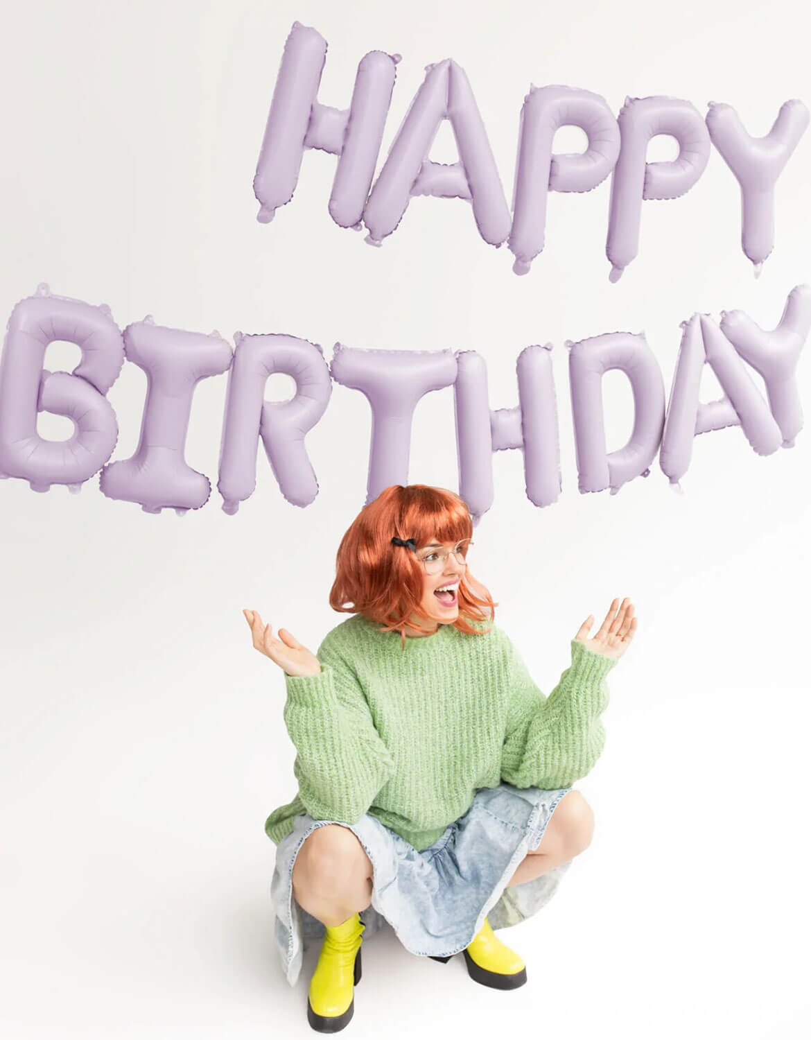 A lady with red hair and green sweater sitting on the floor with Momo Party's 'Happy Birthday' foil balloon set in the background against white cyc.