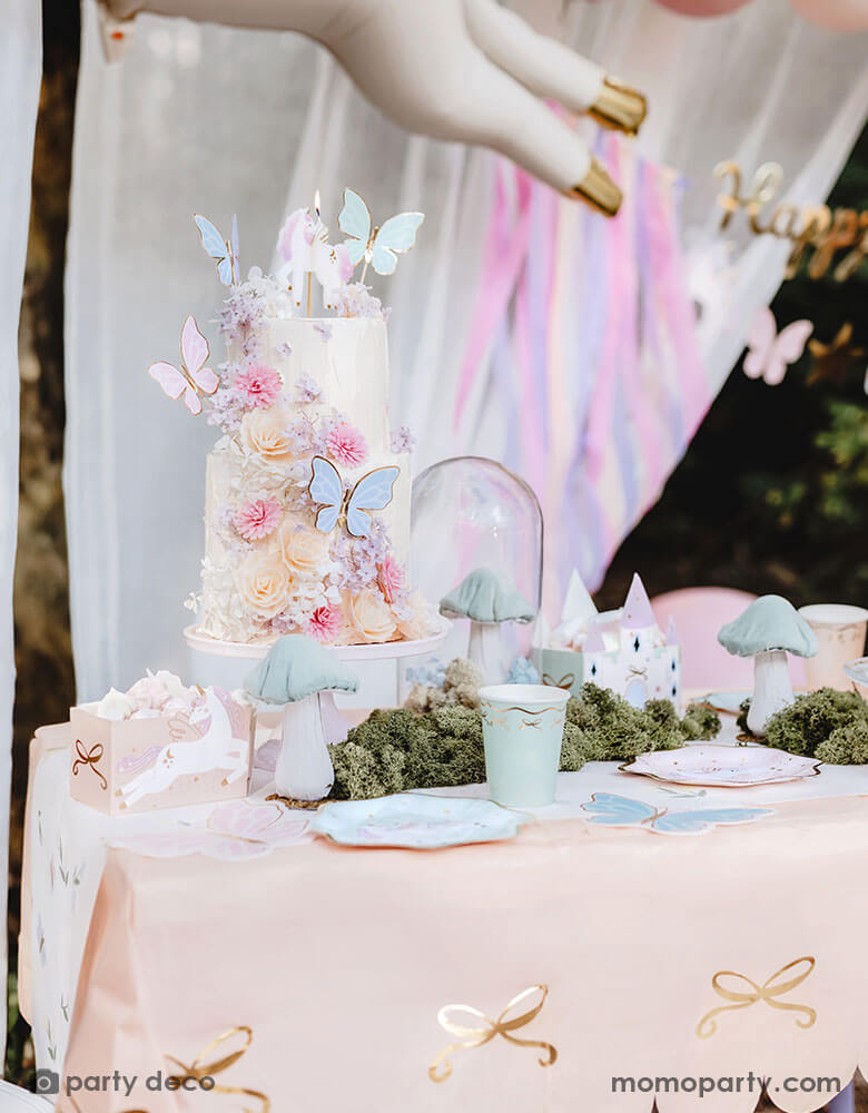 A closeup shot of a whimsical fairy butterfly themed birthday cake with beautiful floral buttercream, topped with Momo Party's pastel butterfly cake toppers. The table is covered with Momo Party's pink bow paper tablecloth with gold foil accent. On the table there are pastel colored tableware including paper plates, party cups, treat boxes and party favor bags. In the middle there's faux grass and mushroom decoration, making this an enchanting set up for girl's magical birthday celebration.