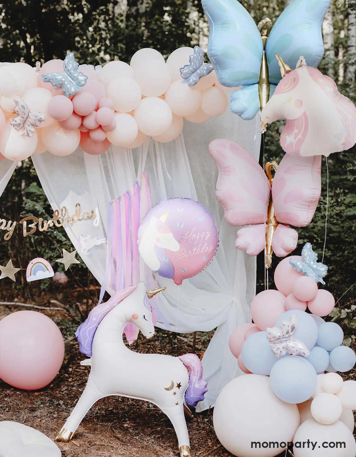 An enchanting unicorn themed birthday party backdrop decoration featuring Momo Party's 37.5" x 34" unicorn standing foil balloon around beautiful pastel colored balloon garlands adorned with butterfly foil balloons, an unicorn balloon and a gold Happy Birthday banner with pastel streamers in the front of a dreamy curtain, making this a whimsical scene for a girly's whimsical unicorn themed birthday party.
