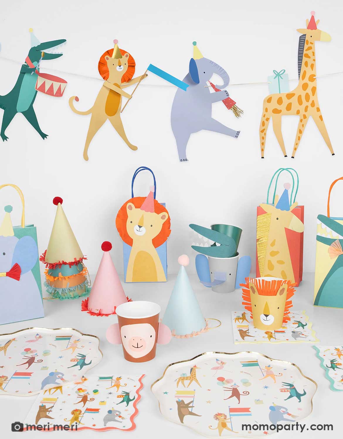 Momo Party's animal parade party collection by Meri Meri featuring colorful animal-themed party supplies including party garland, cups, plates, napkins, hats, and party favor bags with designs of giraffes, elephants, alligators, lions, monkeys and more on a white table. Perfect for young kid's birthday parties.