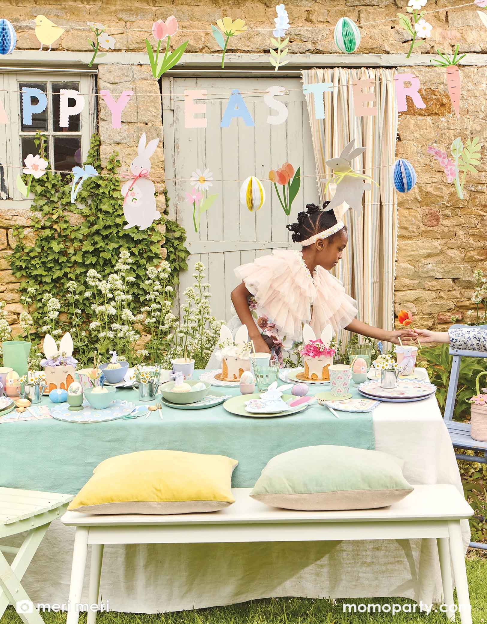 A girl wearing a bunny headband stands next to an Easter party table with Meri Meri's Easter party supplies including the Easter bunny shaped plates, gingham checkered Easter cupcake kit, with pastel aqua blue tablecloth, with spring tulip flowers and Easter eggs and treats on the table. Above the table hung a Happy Easter party garland featuring floral pennants and egg honeycombs, making this a darling setup to celebrate Easter with your kids.