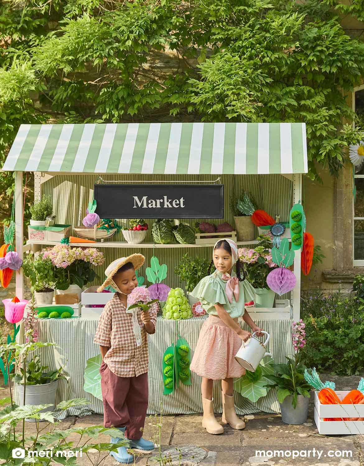 Two children playing at a pretend farmer's market stand with a light green striped canopy that's decorated with Momo Party's giant vegetable honeycomb decorations hanging under the canopy including carrots, beets, lettuces and green pees by Meri Meri. With flowers, plants, produces and various props on the stand. An adorable set up for kid's farmer's market inspired birthday party.