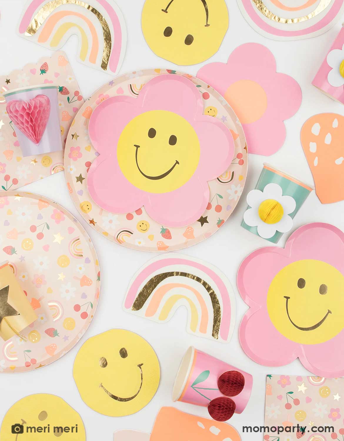 An overshot of a a happy party table featuring colorful party supplies by Meri Meri from Momo Party including flower shaped smiley face plates, flower and rainbow shaped napkins and matching party cups with daisy and heart designs on a white background.