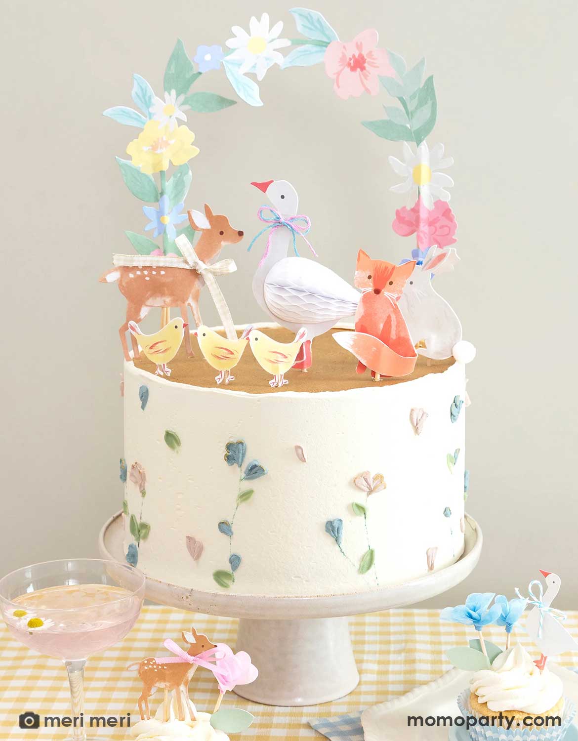 A closeup shot of a rustic cottage themed cake topped with Momo Party's Little Animal cake toppers featuring woodland animals of deer, goose, bunny, fox, chicks and florals by Meri Meri. On the table there are cupcakes decorated with floral little animal cake toppers above the table covered with a soft gingham tablecloth. Making this a perfect set up for a baby shower, an Easter celebration or young kid's storybook rustic cottage inspired birthdays.