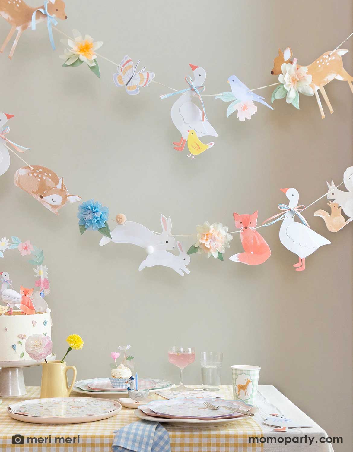 A rustic cottage themed table setting with little woodland animals of deer, goose, bunny, fox, chicks in tablewares and decorations from Momo Party - including party cups, matching plates and napkins and an adorable little animals party garland hung above the table covered with a soft gingham tablecloth. With spring florals in soft yellow and pink on the table, this table is perfect for a baby shower, an Easter celebration or young kid's storybook rustic cottage inspired birthdays.