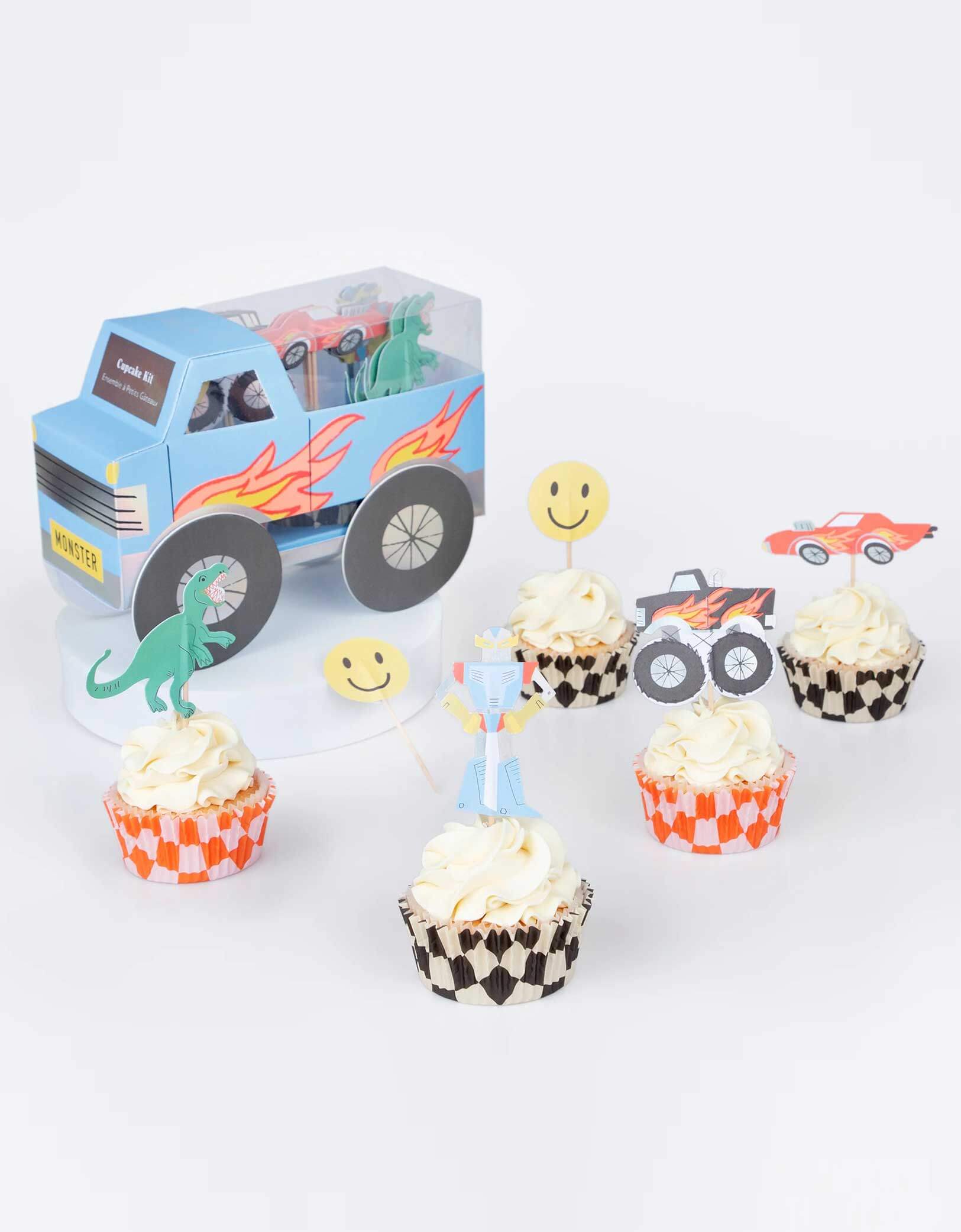 A few cupcakes decorated with Momo Party's Monster Truck Cupcake Kit by Meri Meri, featuring iconic retro toy cupcake toppers, including 5 designs of a monster truck, a car, a robot, a T-rex and a smiley face and checked cupcake cases, to make the most fun and tasty treats. Next to the cupcakes is the monster truck shaped kit box which makes a centerpiece to delight the birthday boy or girl and their guests.