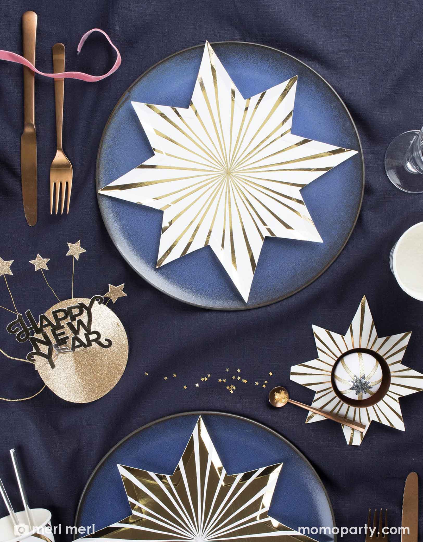 A festive yet modern New Year's party featuring Momo Party's gold starburst shaped plates with foil accent and matching napkins by Meri Meri on a dark blue tablecloth, featuring a gold foil party hat with 'Happy New Year' text.