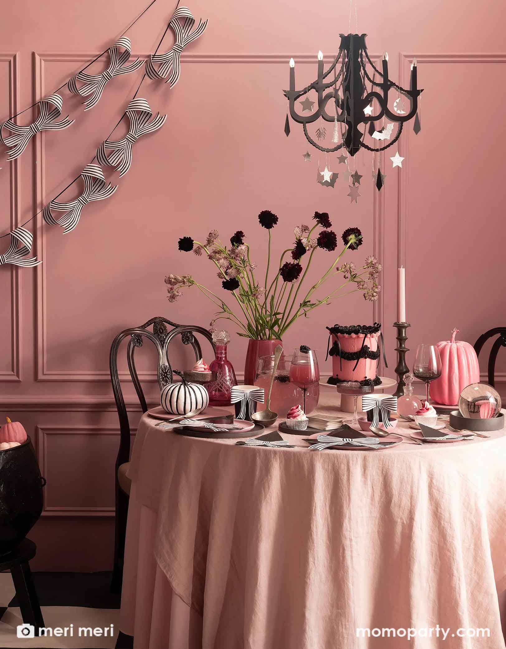 A eerie but elegant pink Halloween party set up featuring Momo Party's black and white stripy bow party tablewares and decorations by Meri Meri including the witch hat shaped plates, napkins, party cups and party garland hung on a pink wall. On the party table, there's a pink and black vintage style buttercream cake adorn with black bows, a pink and a black and white striped pumpkin decorations. With some wine glasses with pink drinks and a pink vase with black poppy flowers and a black chandelier above.