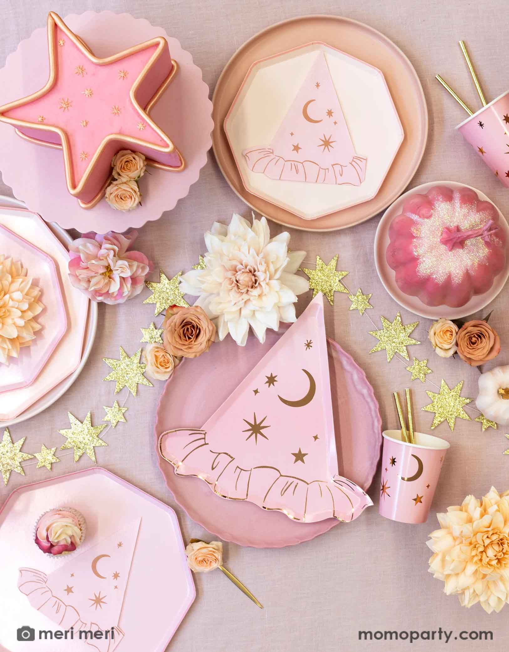 An overhead shot of a pink Halloween party table featuring Momo Party's pink witch themed party supplies with star and moon designs including the pink dinner plates with witch hat shaped plates, napkins and pink party cups with gold foil straws. Around the party tablewares are some dahlia flowers in blush and pale pink, a pink star shaped cake and the gold star glittered garland spread across the table covered with pink tablecloth, making this a chic party look for a non-spooky Halloween celebration!