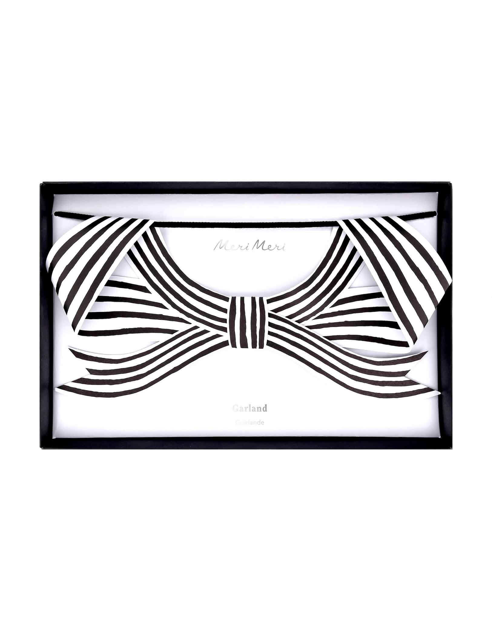 The package of Momo Party's 8 ft Black & White Stripy Bow Garland with bow-shaped ornaments by Meri Meri on a white background. The bold black and white stripes, and soft black velvet ribbon, will give a striking touch indoors or out at your Halloween celebration. Perfect for draping on the party table or mantel, hanging in the porch or windows, or swaying from trees. Plus, it's reusable to bring effortless elegance, and a hint of eerie charm, year after year.