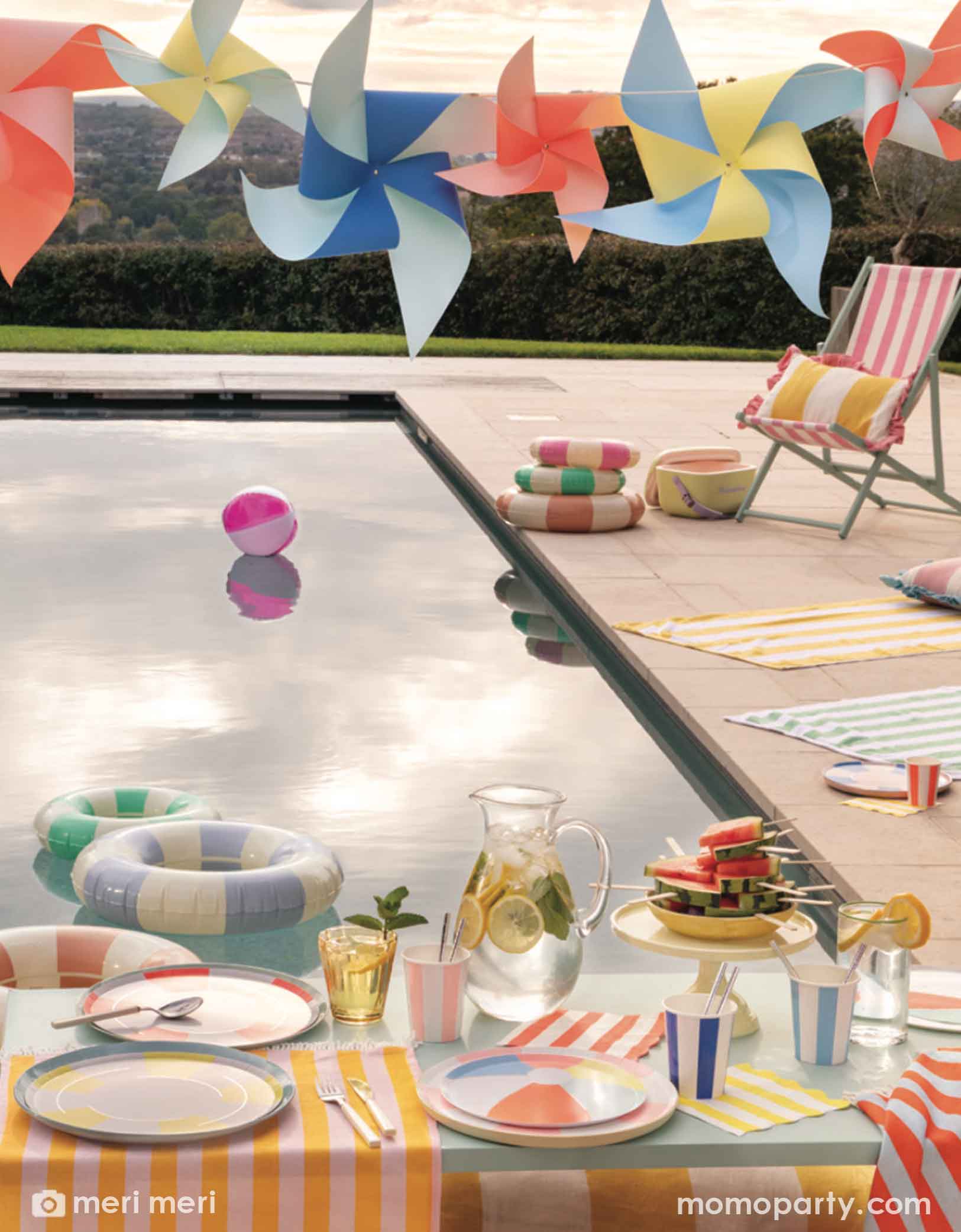 A poolside party setup featuring colorful beach towel striped themed tablewares from Momo Party by Meri Meri including beach ball shaped plates, striped napkins, party cups, party garlands decorations, pool floats, and a table with summer food and drinks.