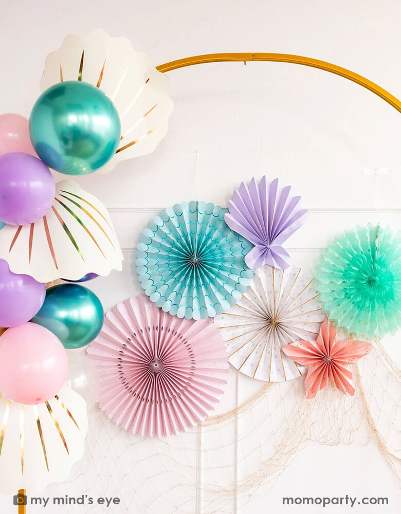 An elegant white wall decorated with mermaid inspired party decorations from Momo Party including under the sea paper fan set in pastel colors colors of aqua, pink, lilac, teal, cream and peach, along with mermaid themed party balloons in coordinating colors with My Mind's Eye's large cream shell shaped paper decorations, with a fishing net hung underneath, this makes a perfect backdrop for an enchanting kid's under the sea, mermaid birthday party.