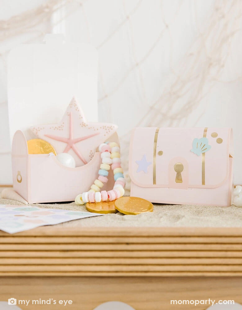 Two under the sea treasure chest box shaped treat boxes from Momo Party on a wooden party table. These light pink treat boxes are decorated with adorable pastel colored seashell and sea star stickers. In the treat boxes there's a pink sea star shaped sugar cookie, gold chocolate coins and a candy necklace, making this a great party favor idea for a kid's mermaid or under the sea themed birthday party.