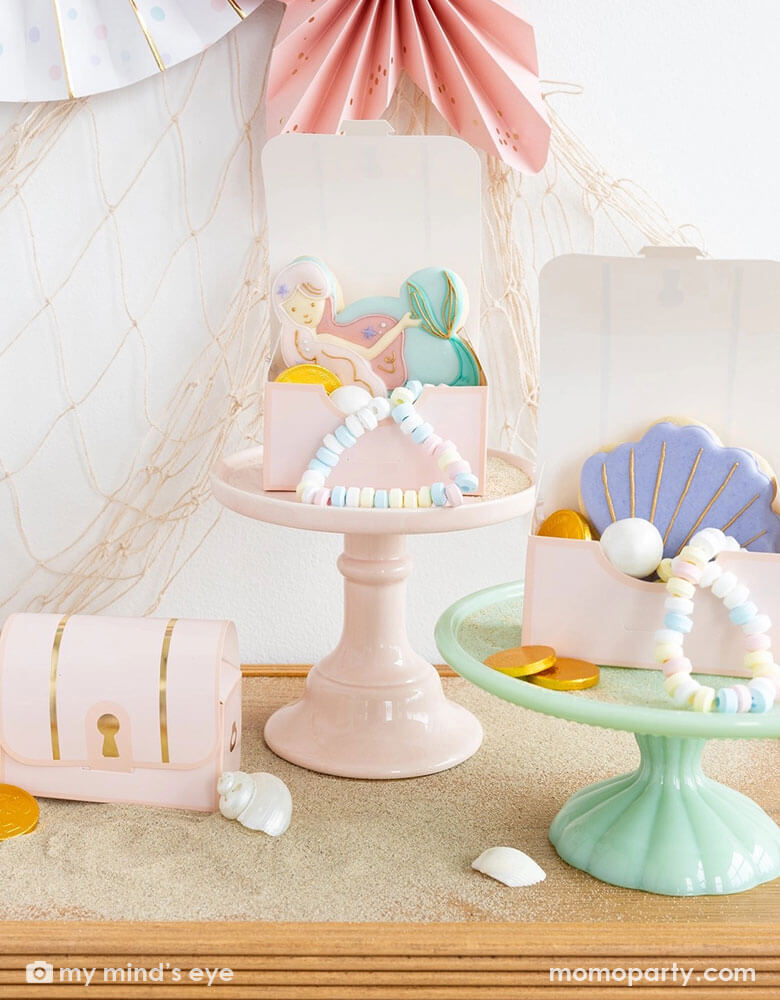 A closeup shot of an adorable mermaid party table decorated with Momo Party's pink under the sea treasure chest treat boxes on darling pastel cake stands. In the treat boxes are pastel candy necklaces, some gold chocolate coins, mermaid and seashell sugar cookies in coordinating pastel colors. In the back of the wall there's a fishing net decoration draped from the wall, making this a great decorating inspo for kid's mermaid/under the sea birthday parties.