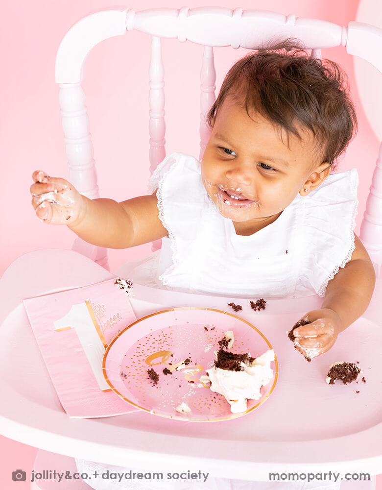 A baby girl sitting on a high chair, eating cakes with the Jollity & co. Milestone Pink Onederland plates and Guest Napkins to celebrate her 1st birthday party