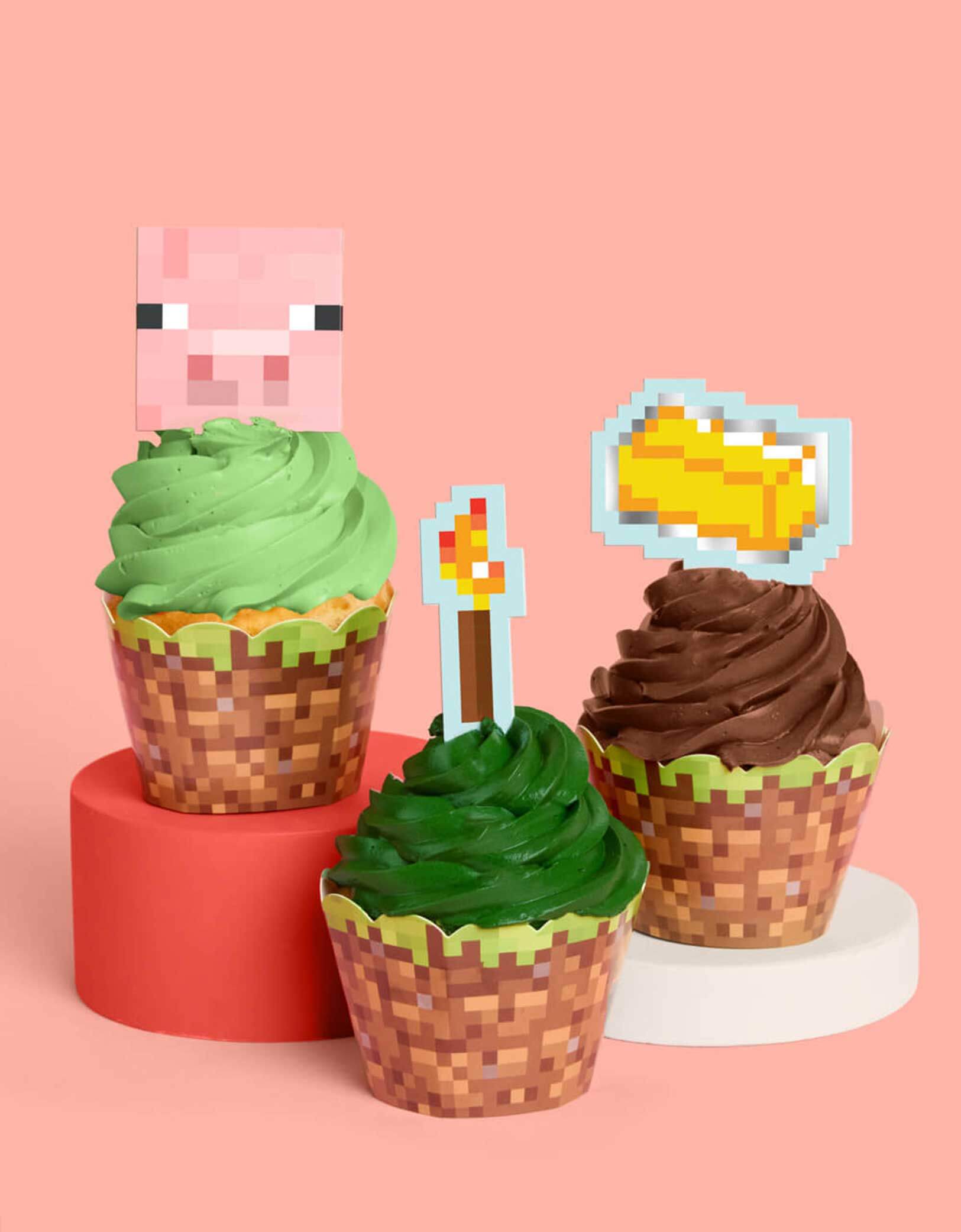 Three cupcakes with green and brown frosting with decorative Minecraft themed toppers and liners featuring a pig face, a pixel torch, and a gold brick from Momo Party on a pink background.