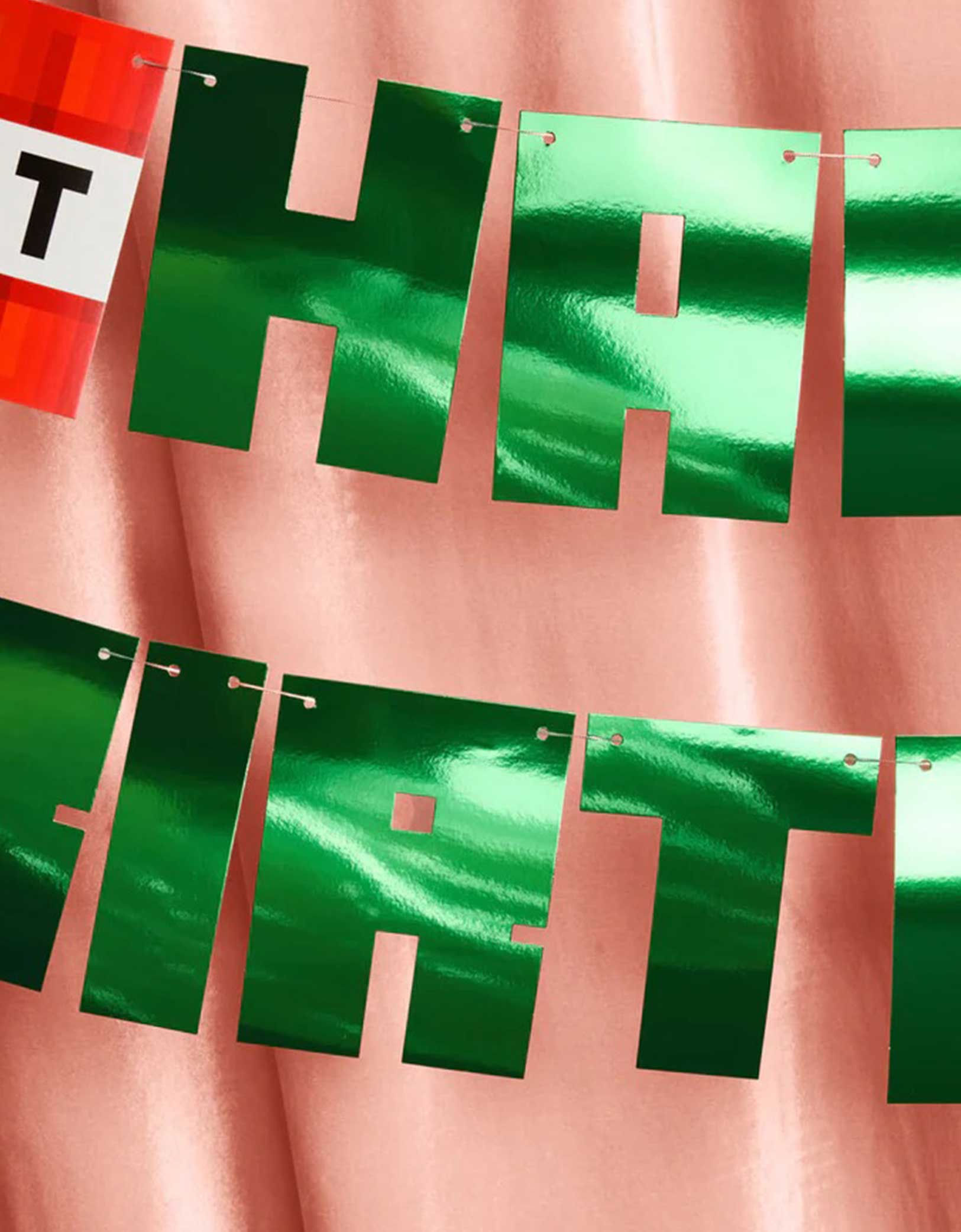 A closeup shot of Momo Party's Minecraft Pixel Birthday Banner with pixelated pennants including a TNT block pennant and green foiled Happy Birthday pennants on a pink curtain background.