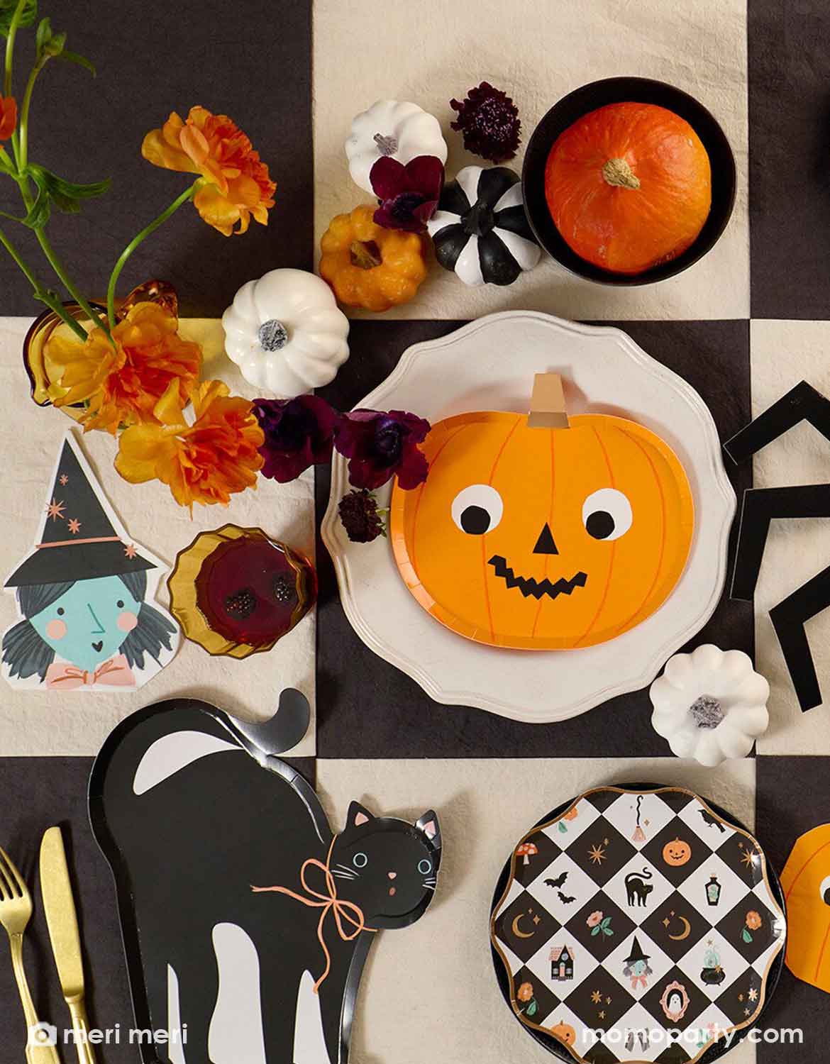 A styled Halloween table setting featuring Momo Party's Halloween themed party supplies including a pumpkin shaped plate with goggly eyes, a black cat shaped plate, a which shaped napkin and a black and white checkered pattern dessert plate on a black checkered tablecloth. Along with orange and white flowers, small pumpkin decorations in black, white and orange - making this a perfect look for a modern Halloween tablescape.