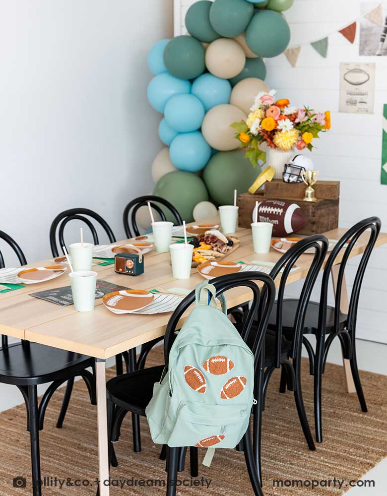 A kid's football birthday party set up with a modern party table filled with football themed tableware from Momo Party. In the center of the table there's a cheese charcuterie board and some decorations like a football, a trophy and a football helmet as the centerpiece. On one of the chairs, there's a backpack with football patches on it. On the wall there are some old school football party banners, posters and party balloons to add some festivities to the celebration!