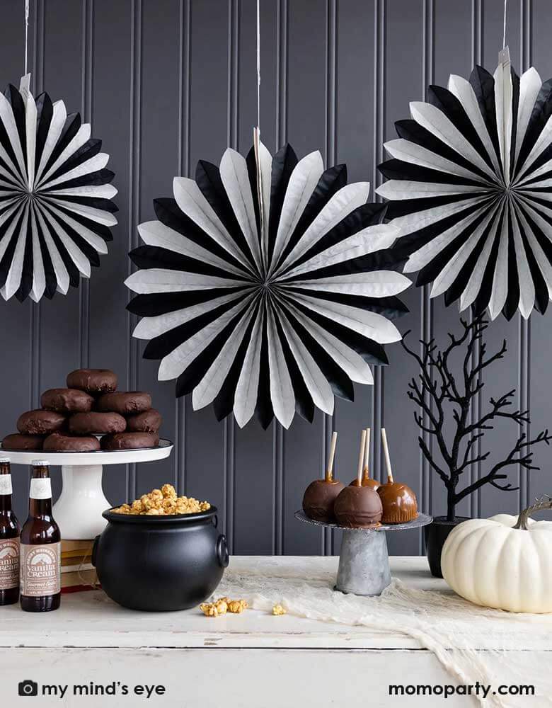 A classic black and white Halloween table decorated with white pumpkins, creepy tree, caramel apples and a witch cauldron filled with popcorn. In the back the wall was decorated with My Mind's Eye's oversized black and white tissue paper fans. All makes a simply yet iconic Halloween decorating inspo this Halloween season.