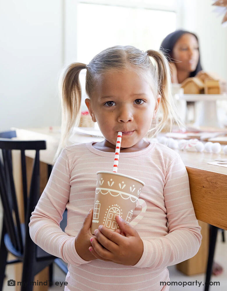 In pink-striped PJs, a girl savors a sip through Gingerbread Reusable Straws, cradling Gingerbread House Paper Party Cups with Handles. In front of a wooden dinner table, she's surrounded by captivating gingerbread-themed party essentials, all available at momoparty.com